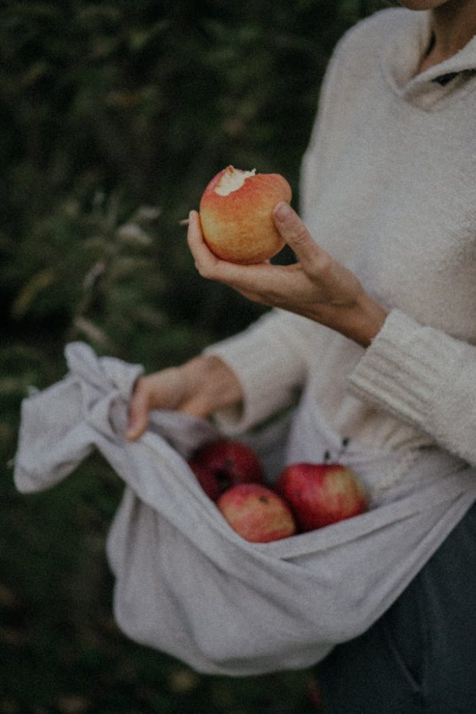 Close-up of a woman in a white sweater holding and eating apples outdoors in fall.